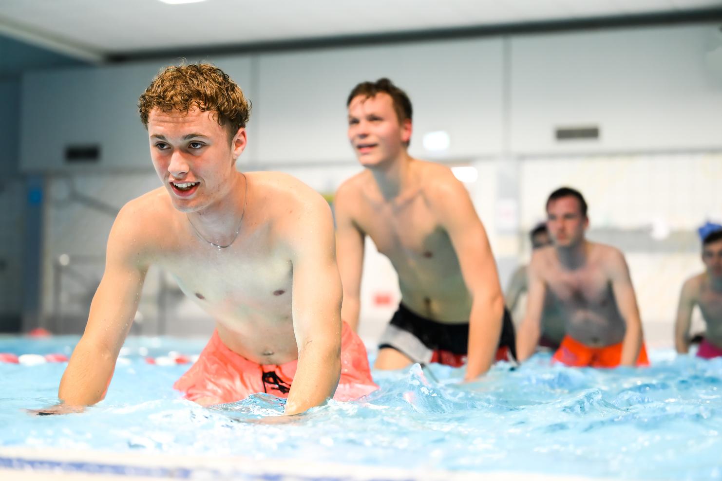 Vier junge Männer in Badebekleidung üben im Wasser eines Schwimmbeckens, einige lachen und spritzen.